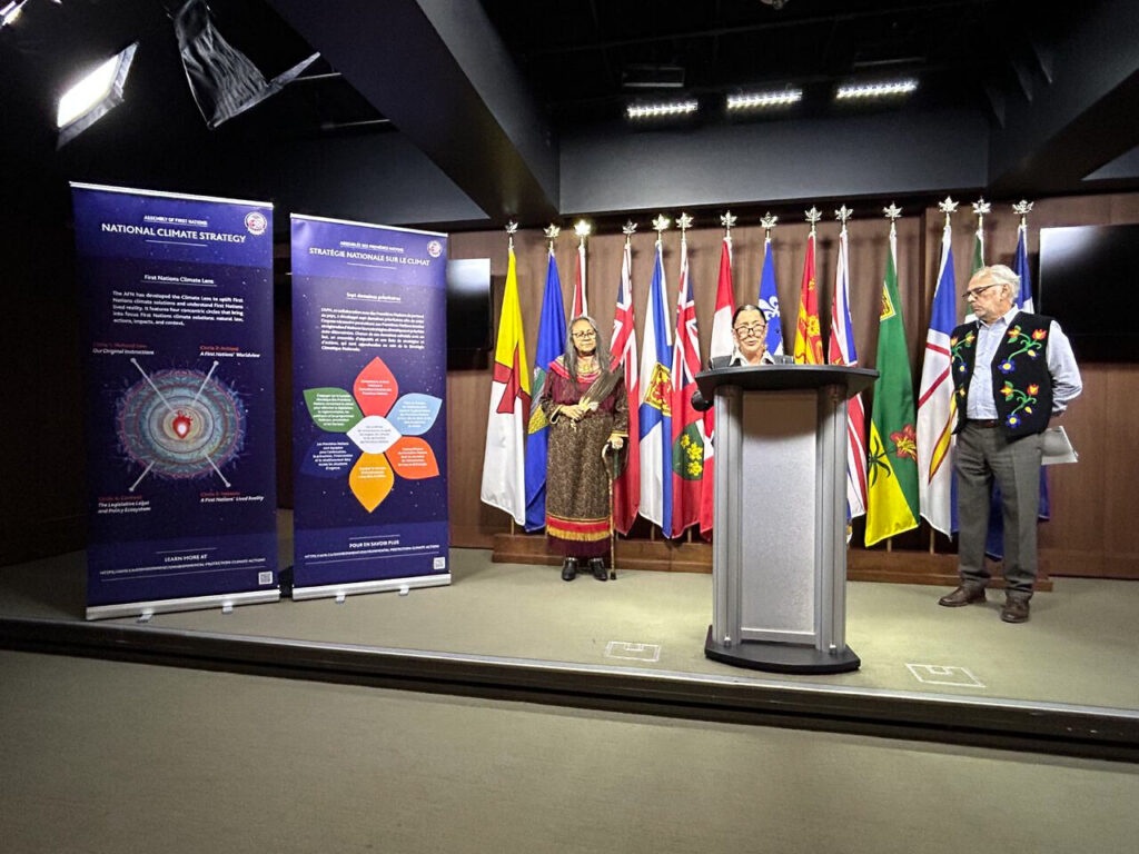 Photo taken from Climate Gathering Forum in 2023, displaying Climate Gathering Pull-Up Banner with Canadian Knowledge Keeper talking at Podium