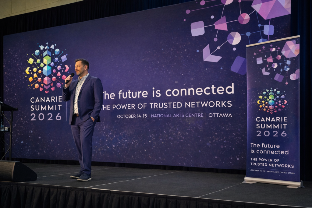 Photograph of a conference stage featuring CANARIE Summit 2026 branding. A man in a navy suit stands on stage holding a microphone, speaking to the audience. Behind him is a full-width printed backdrop in deep blue with colourful geometric network graphics and the text: “CANARIE Summit 2026 – The future is connected: The power of Trusted Networks. October 14–15 | National Arts Centre | Ottawa.” A matching branded pull-up banner stands to the right of the stage, mirroring the summit logo, tagline, and event details. Dark stage curtains frame the backdrop.